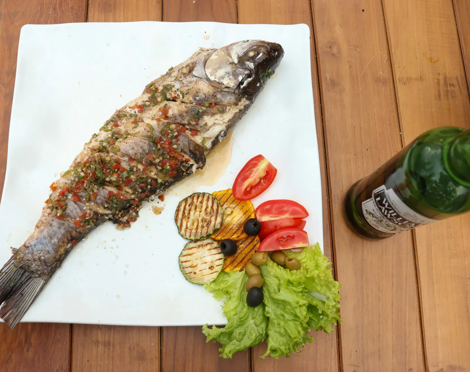 A grilled fish placed on a white plate next to a drink on a wooden table.
